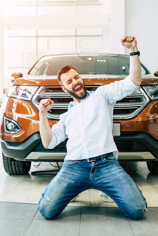 Glücklicher Besitzer eines neuen Autos. Emotionaler gutaussehender Mann in lässiger Kleidung steht glücklich auf den Knien, wenn er ein Auto im Autohaus kauft